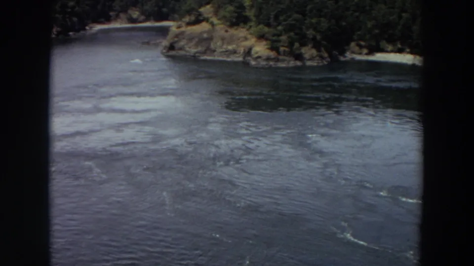 1964 Washington 8mm frame with a wide dark waterway, forested bank, rocky outcrop, and small pale beach in view.
