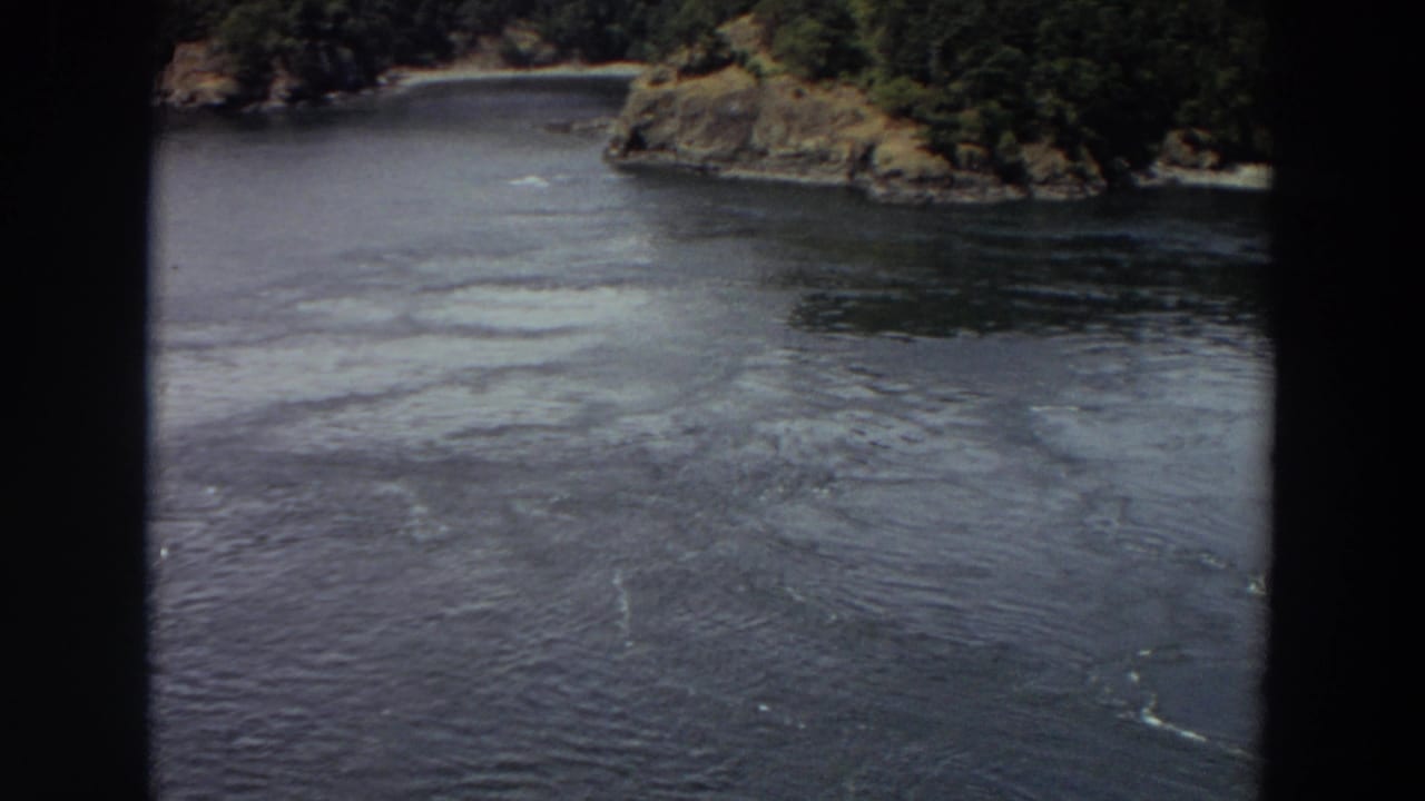 River surface with pale swirling current below a wooded rocky shoreline.
