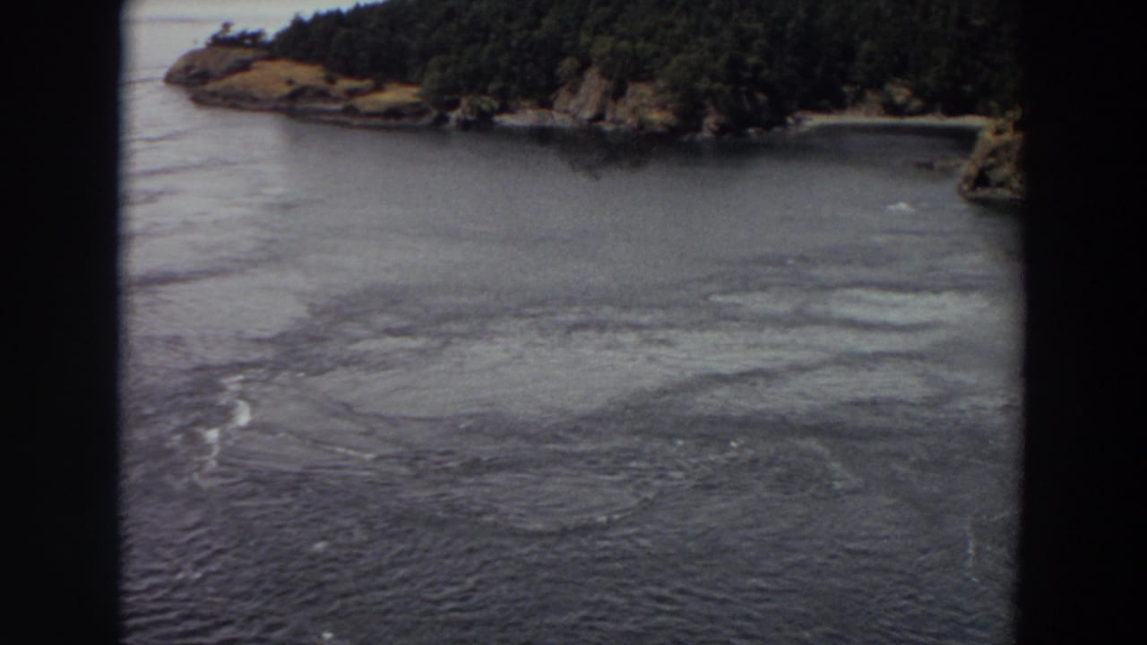 1964 Washington 8mm frame showing a river channel, circular surface swirls, and a wooded rocky shore under muted light.