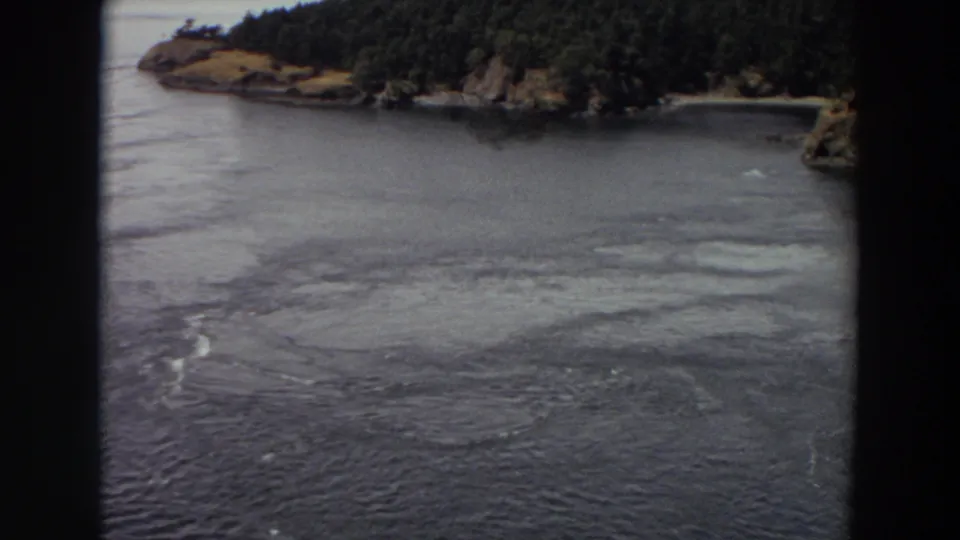 1964 Washington 8mm frame showing a river channel, circular surface swirls, and a wooded rocky shore under muted light.