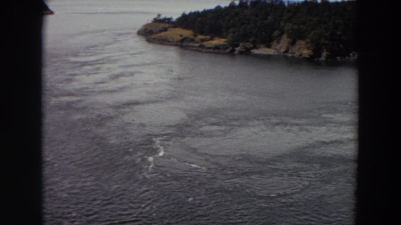1964 Washington 8mm frame capturing rippled river water, pale swirling current, and a tree-covered shoreline beyond.