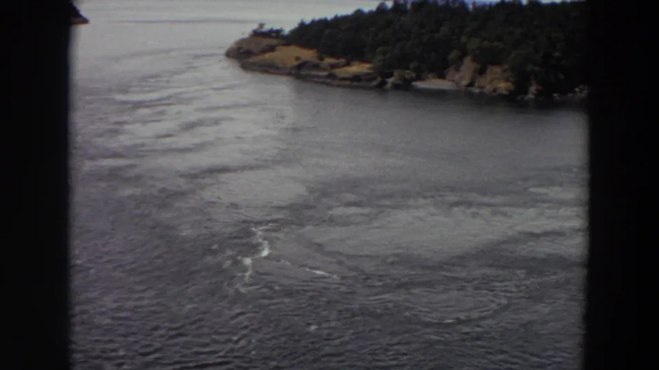 1964 Washington 8mm frame capturing rippled river water, pale swirling current, and a tree-covered shoreline beyond.