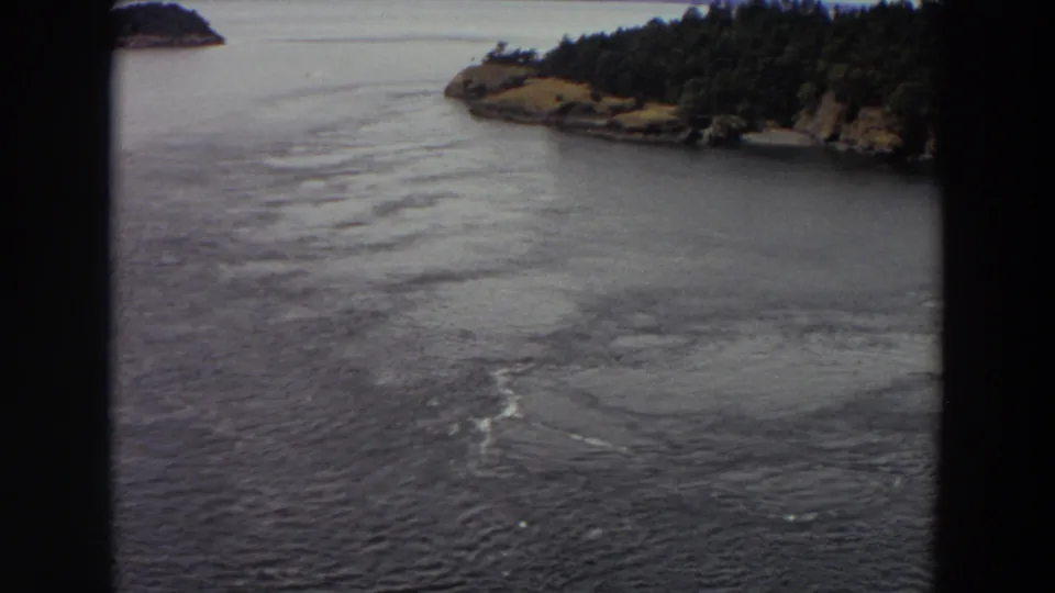 1964 Washington 8mm frame showing rippled water, a tree-lined shoreline, and a small island, partially framed by dark foreground borders.