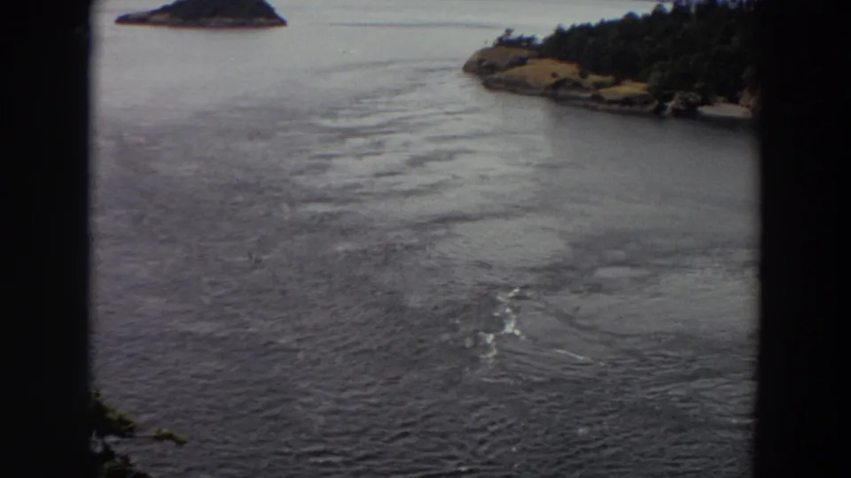 1964 Washington home-movie frame of calm rippled water with a wooded shore at right and a small island in the distance.