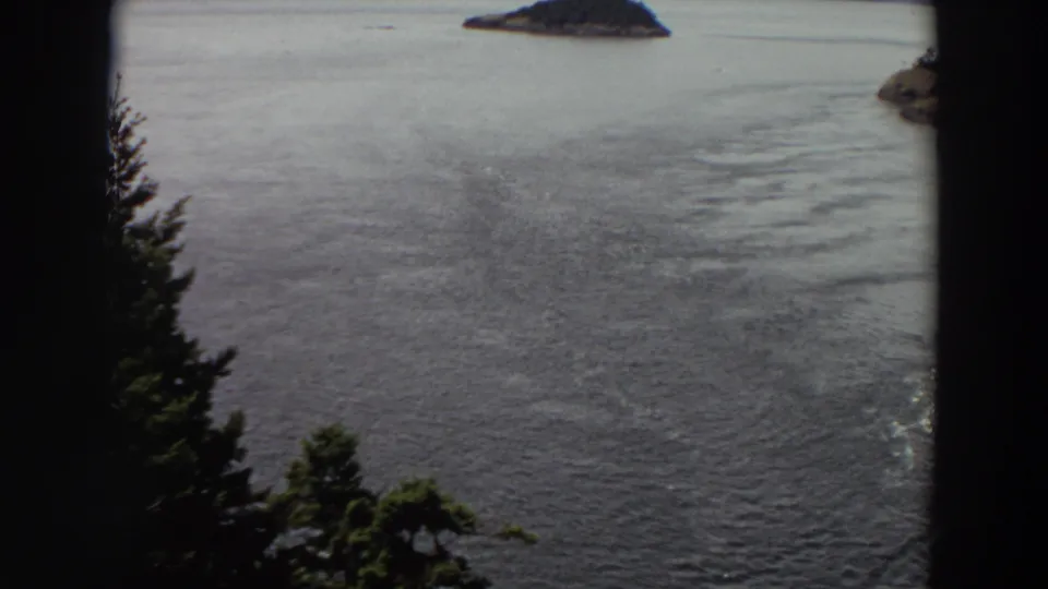 1964 Washington home-movie frame of open water, a small island near the horizon, and a partial wooded shoreline at right.