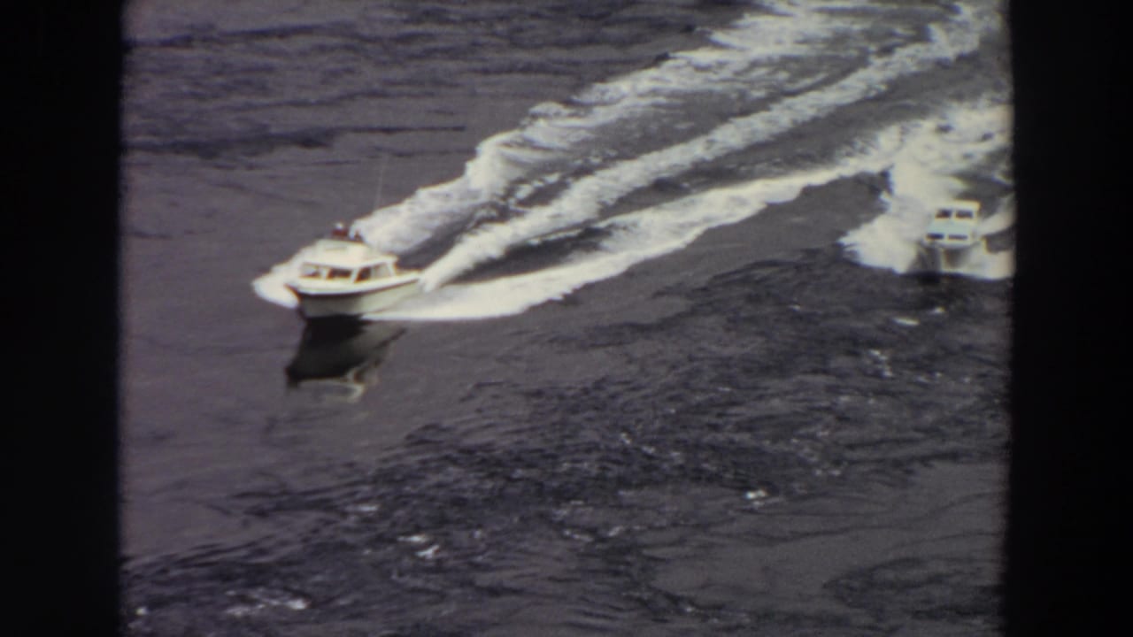 1964 Washington home-movie frame with two speedboats crossing the water, their wakes forming bright arcs behind them.