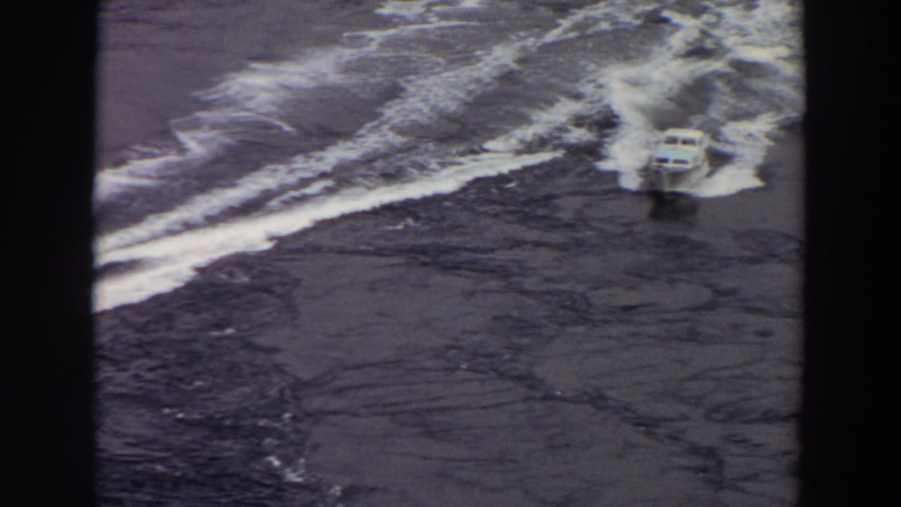 1964 8mm frame of a small white motorboat approaching through curved wake patterns on calm Washington water.