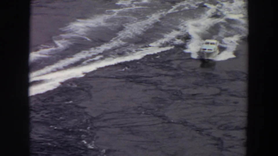 1964 8mm frame of a small white motorboat approaching through curved wake patterns on calm Washington water.