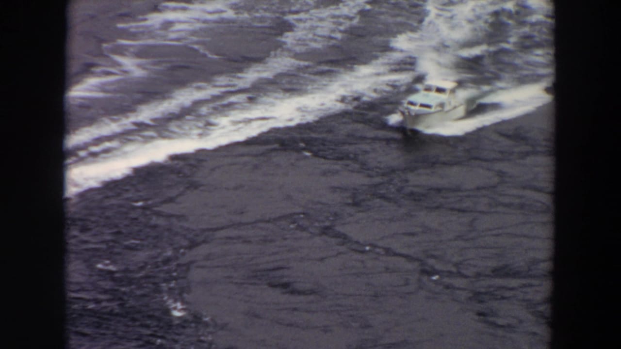 High-angle 1964 Washington home-movie frame showing a motorboat crossing dark water with a bright wake behind it.