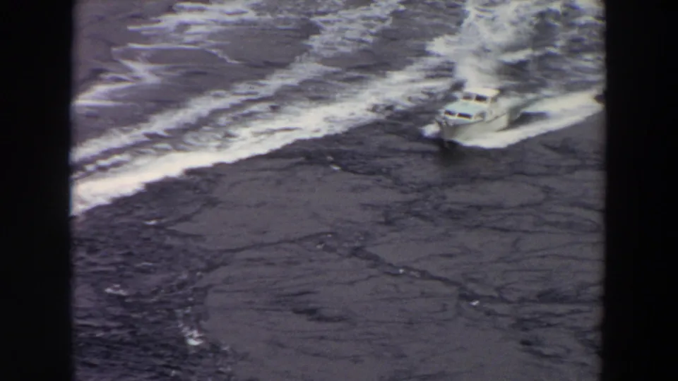 High-angle 1964 Washington home-movie frame showing a motorboat crossing dark water with a bright wake behind it.