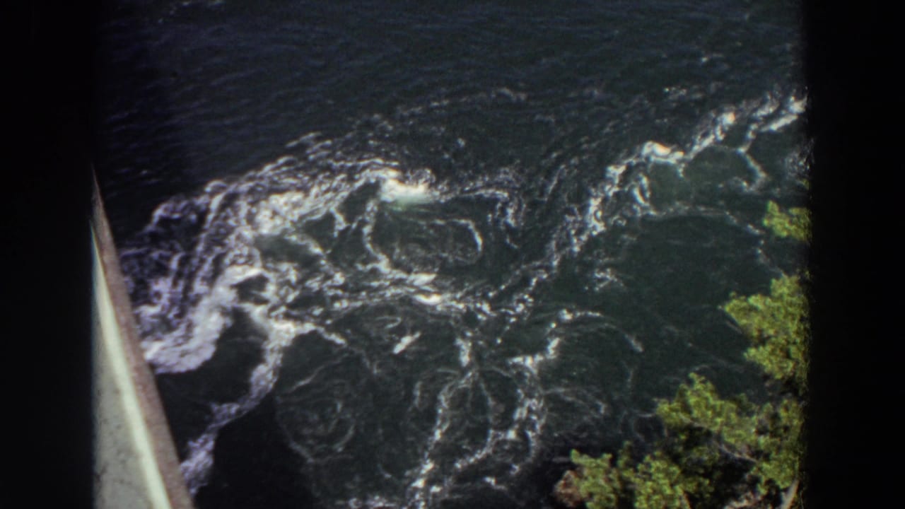 Fast river water with white foam, green foliage, and a bridge or railing edge at left.