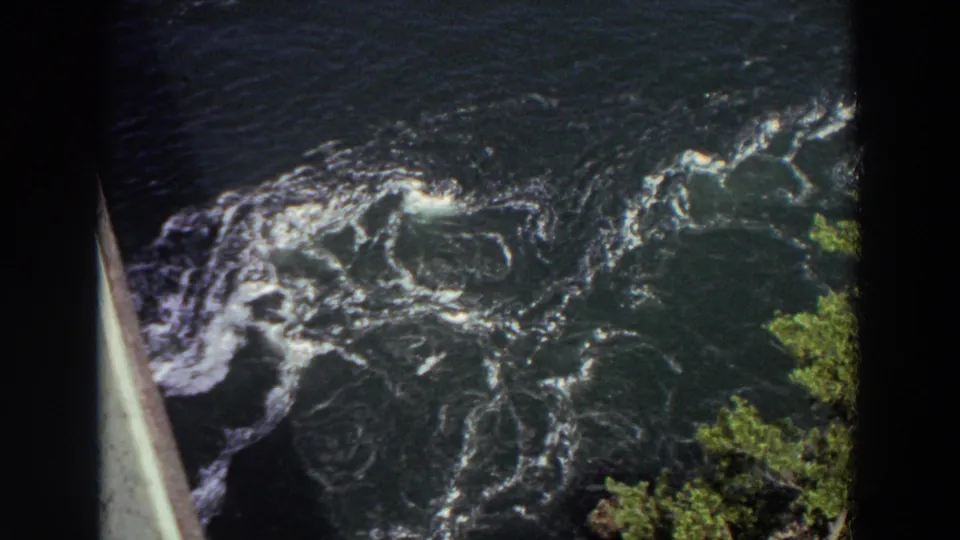 1964 Washington 8mm frame of turbulent river water with whitecaps, eddies, green bank foliage, and a bridge or railing edge visible at left.