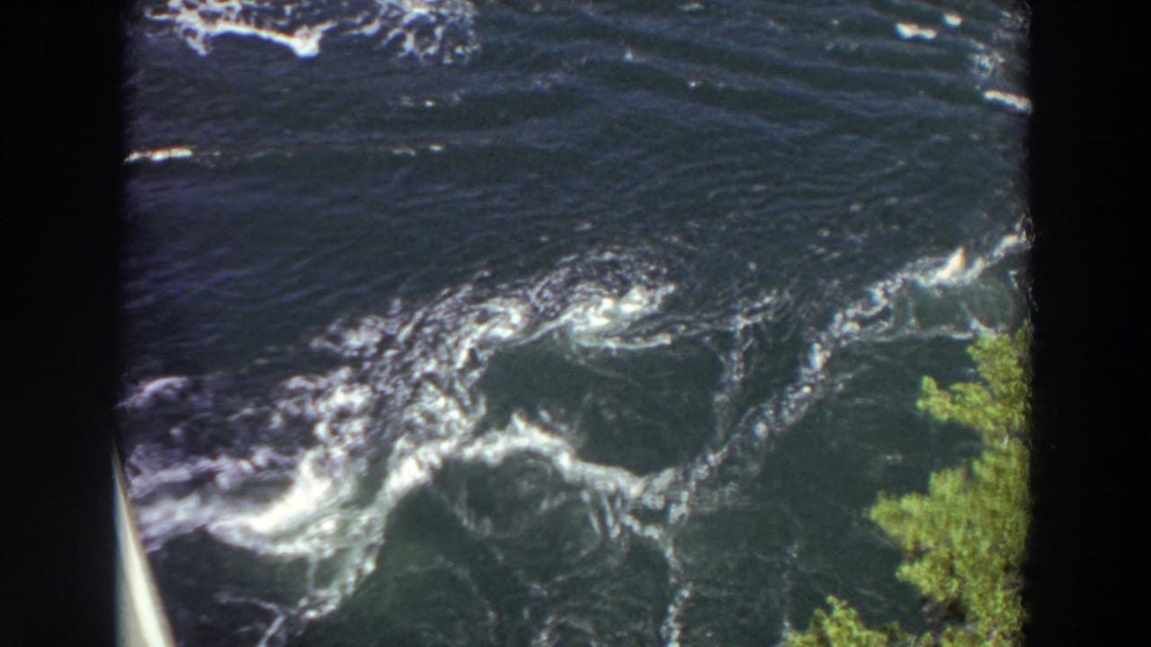 1964 Washington 8mm frame of fast-moving river water with foam patterns, visible eddies, green foliage, and a pale bridge or railing edge.