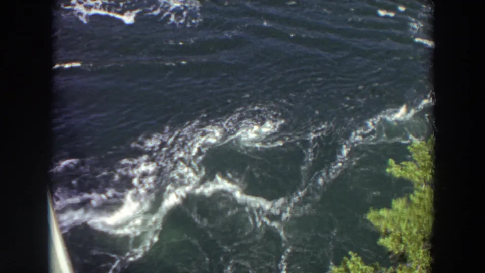 1964 Washington 8mm frame of fast-moving river water with foam patterns, visible eddies, green foliage, and a pale bridge or railing edge.