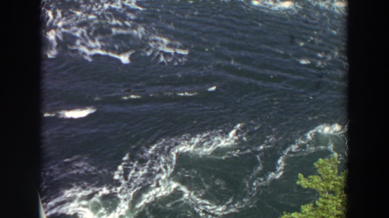 1964 Washington frame showing turbulent dark river water, streaks of white foam, circular current patterns, and foliage at the riverbank.