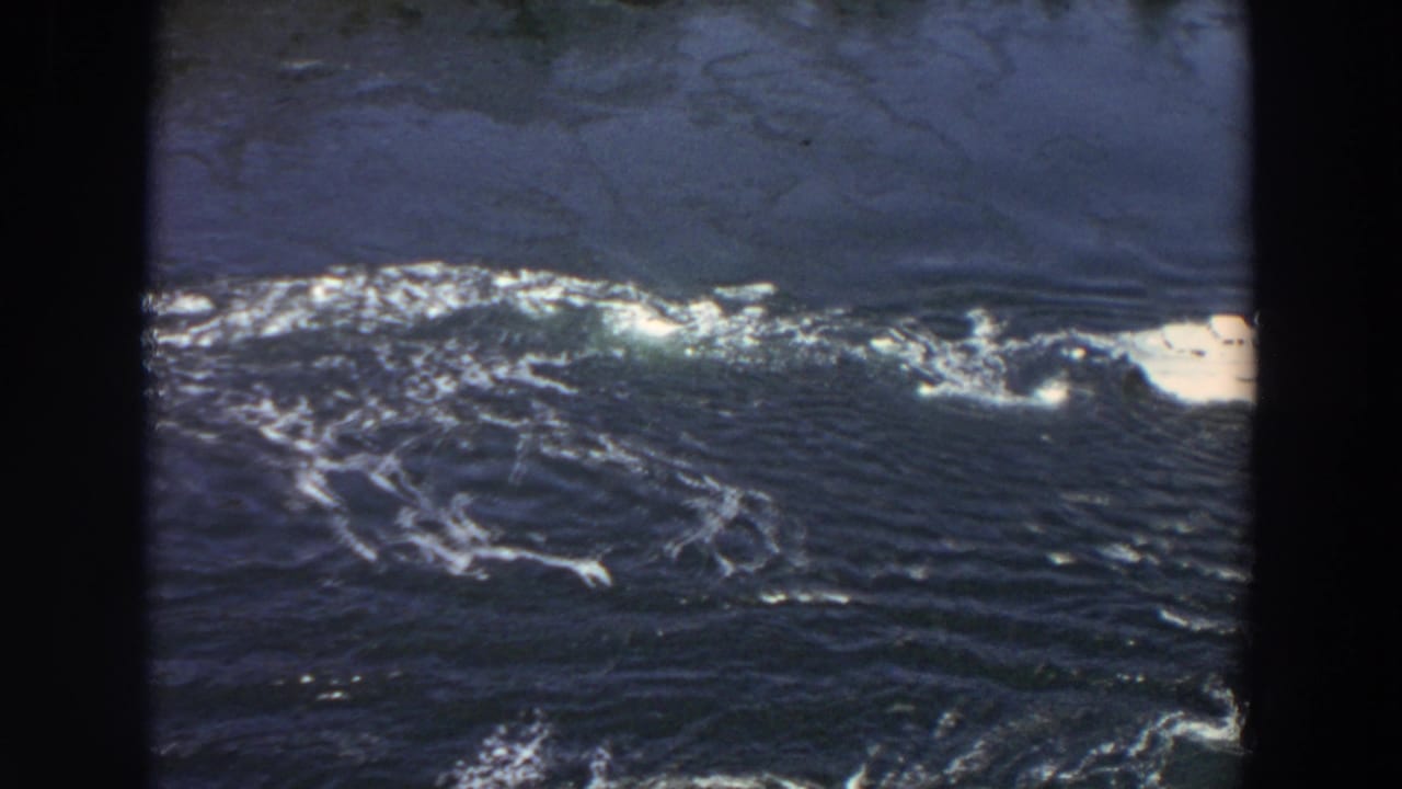 1964 Washington 8mm frame with dark river current, white foam streaks, and a pale shoreline or water edge at the right side.