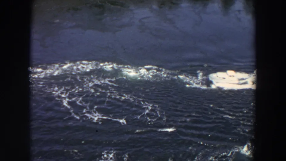 A white yacht moves across dark blue water in 1964 Washington home-movie footage, with shoreline and film-frame borders visible.