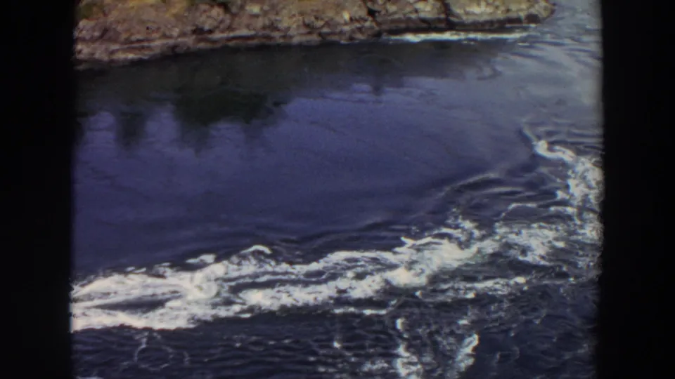 1964 Washington 8mm frame showing foamy water currents beside a rocky shoreline and dark lake surface.