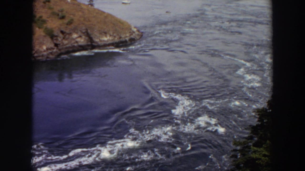 1964 Washington 8mm view of a rocky point, churning outlet water, calmer lake surface, and small distant boats.