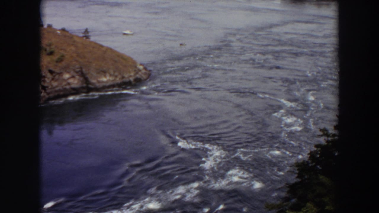 1964 Washington frame showing foamy outlet currents, a rocky shoreline, open lake water, distant boats, and trees at the lower edge.