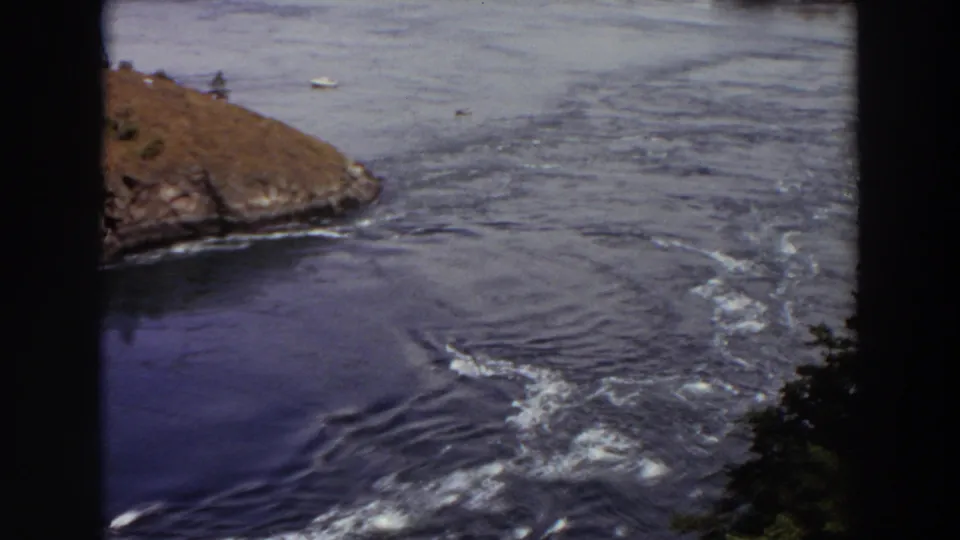1964 Washington frame showing foamy outlet currents, a rocky shoreline, open lake water, distant boats, and trees at the lower edge.