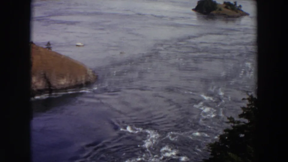 1964 Washington 8mm view of swift water moving around rocky shorelines and small tree-covered islands.