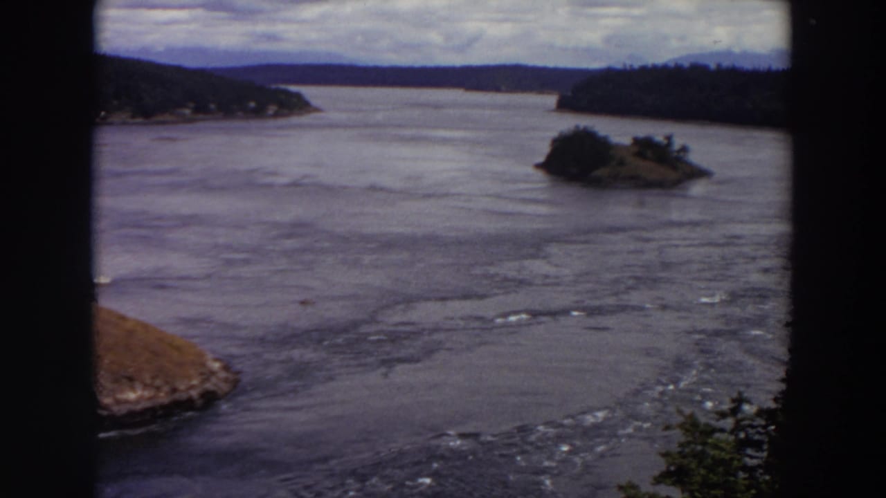 1964 Washington home-movie frame showing a forested island, rocky shoreline, and moving water under cloudy sky.