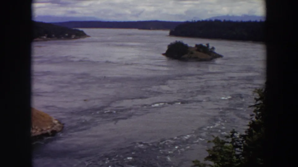 1964 Washington 8mm frame showing rippled water, a small tree-covered island, distant wooded hills, and dark frame edges.