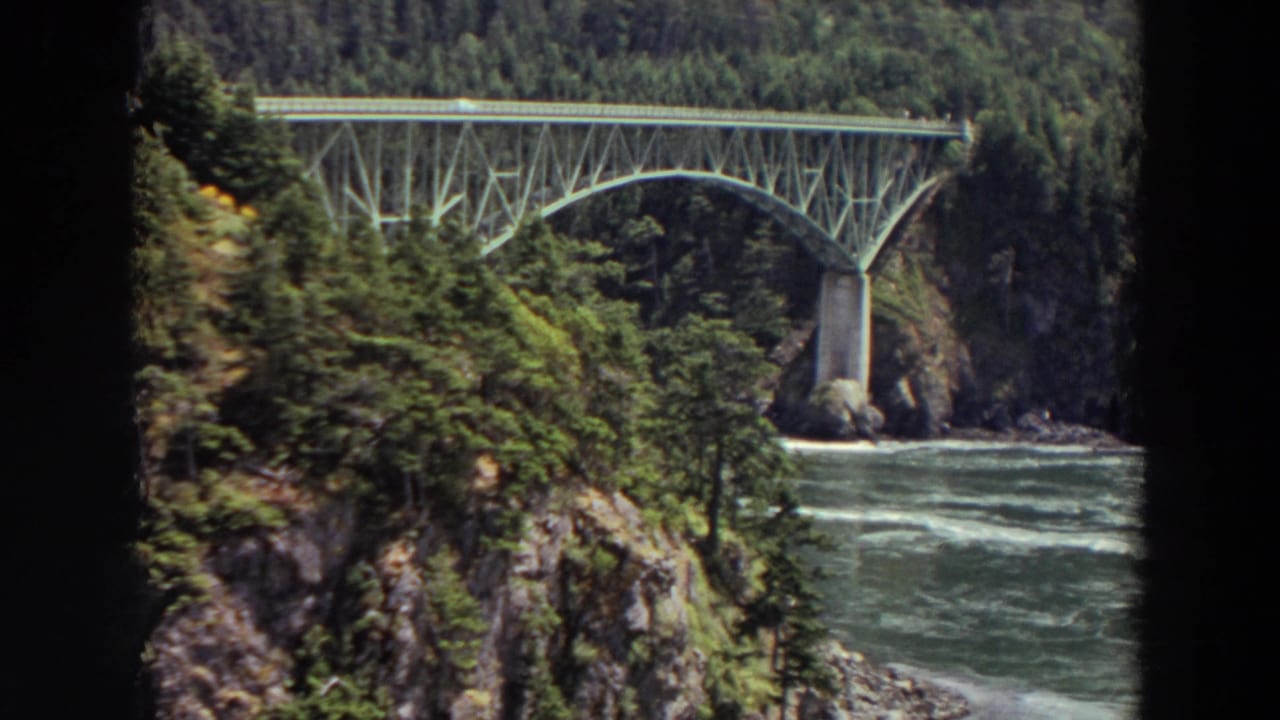 Steel arch bridge over a river gorge with evergreen forest and rocky cliffs.