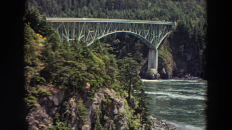 1964 Washington 8mm frame showing a steel arch bridge spanning a rushing river amid evergreen forest and cliffs.