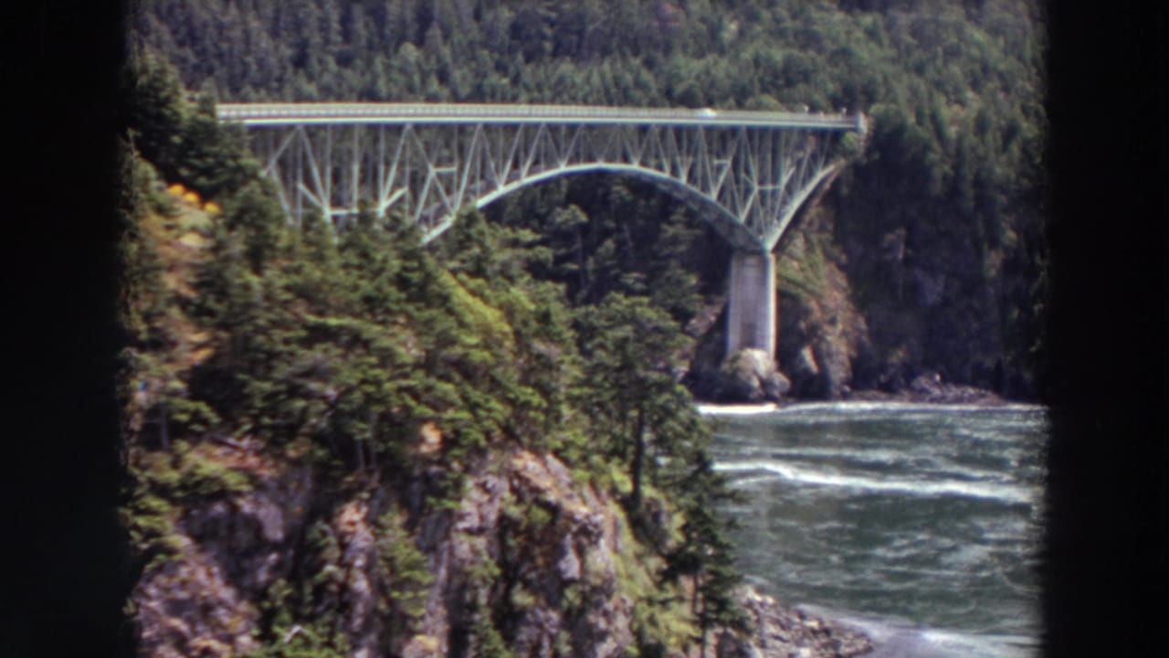 Steel arch bridge over a fast river, framed by evergreen forest and rocky canyon slopes.