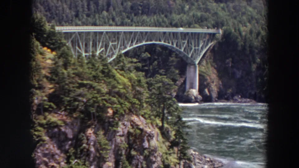 1964 Washington 8mm frame showing a steel arch bridge spanning moving water between forested rocky cliffs.