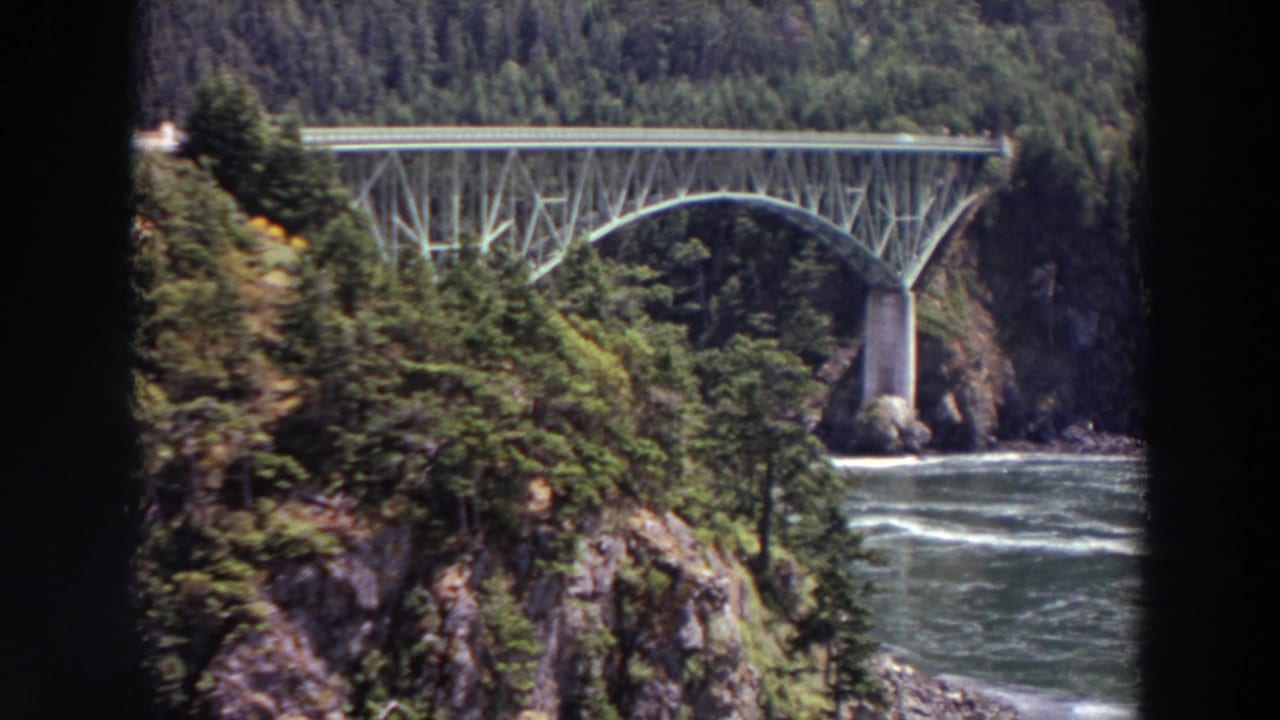 1964 Washington travel footage frame with a steel bridge, dense evergreens, rocky canyon walls, and moving river water below.