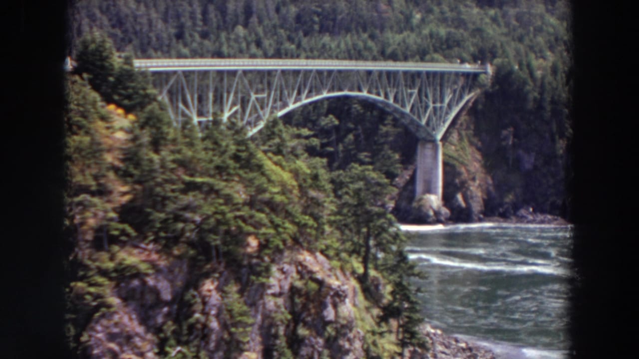 1964 Washington 8mm still showing a steel arch bridge seen past trees and rocky slopes above a fast-moving river.