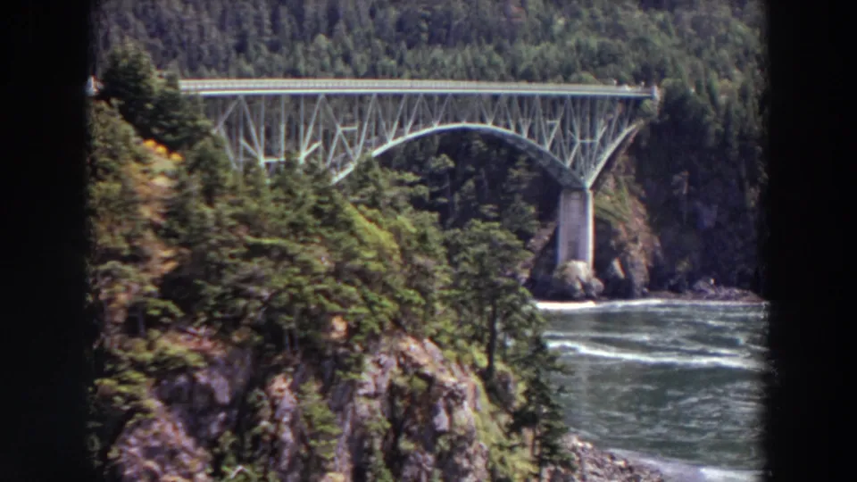 1964 Washington 8mm still showing a steel arch bridge seen past trees and rocky slopes above a fast-moving river.