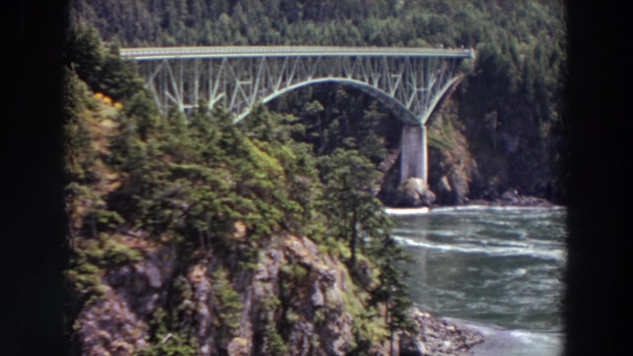Steel arch bridge crossing water between wooded rocky slopes.