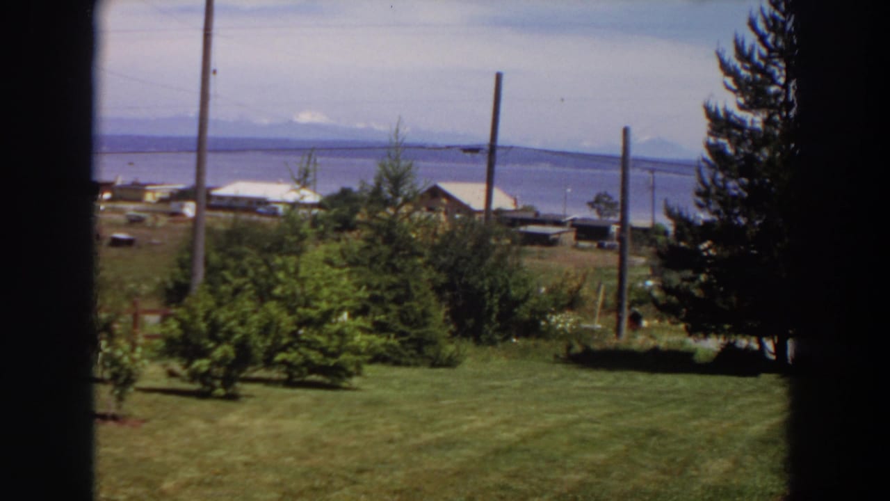 1964 Washington 8mm frame showing a rural yard and waterfront buildings with mountains across the water.