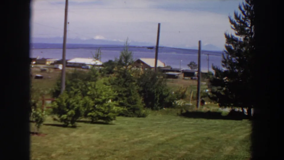 1964 Washington 8mm frame showing a rural yard and waterfront buildings with mountains across the water.