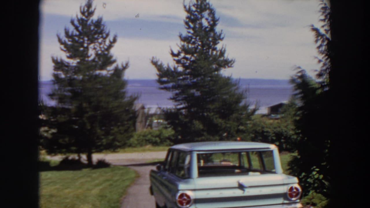 1964 Washington home-movie frame with the rear of a pale station wagon, residential greenery, a road, water, and distant hills.
