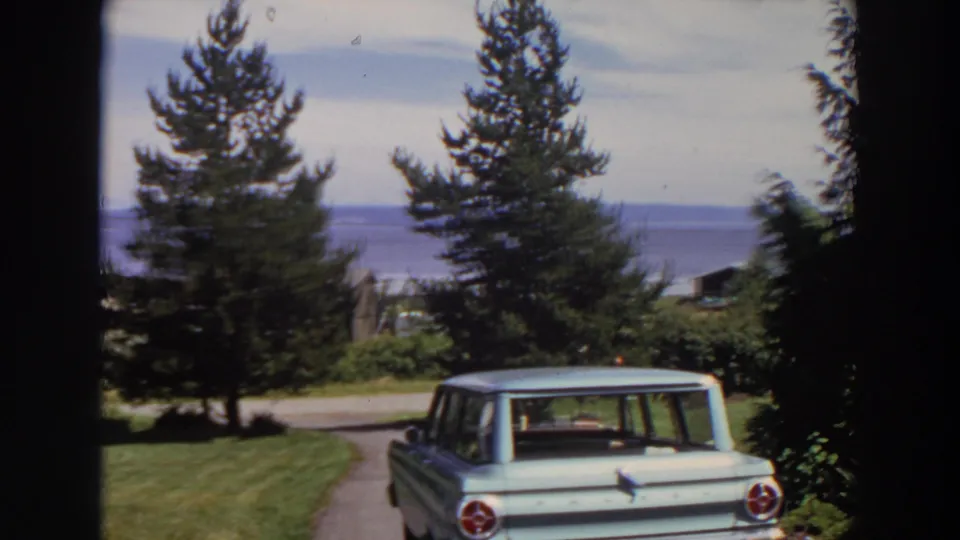 1964 Washington home-movie frame with the rear of a pale station wagon, residential greenery, a road, water, and distant hills.