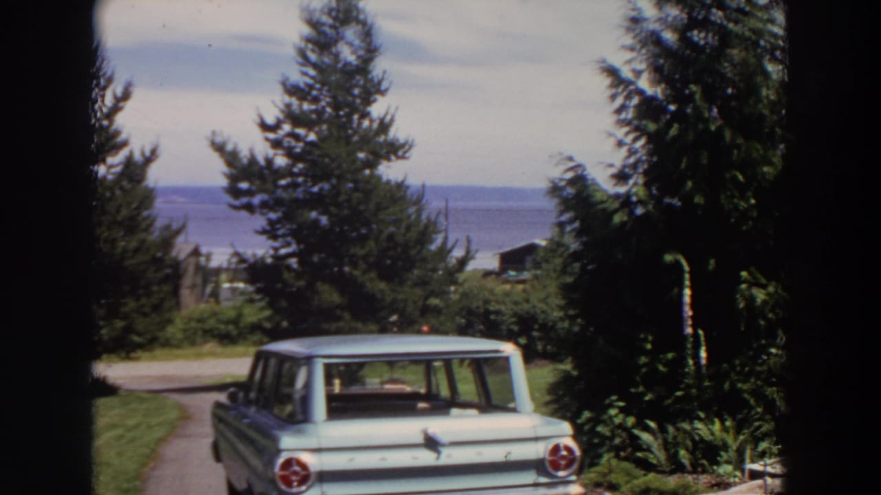 Washington 1964 8mm frame showing a parked station wagon framed by lawn, tall trees, roadside, and a blue coastal vista.