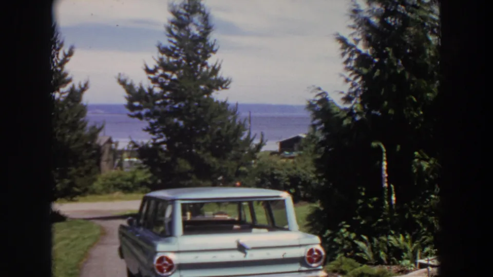 Washington 1964 8mm frame showing a parked station wagon framed by lawn, tall trees, roadside, and a blue coastal vista.