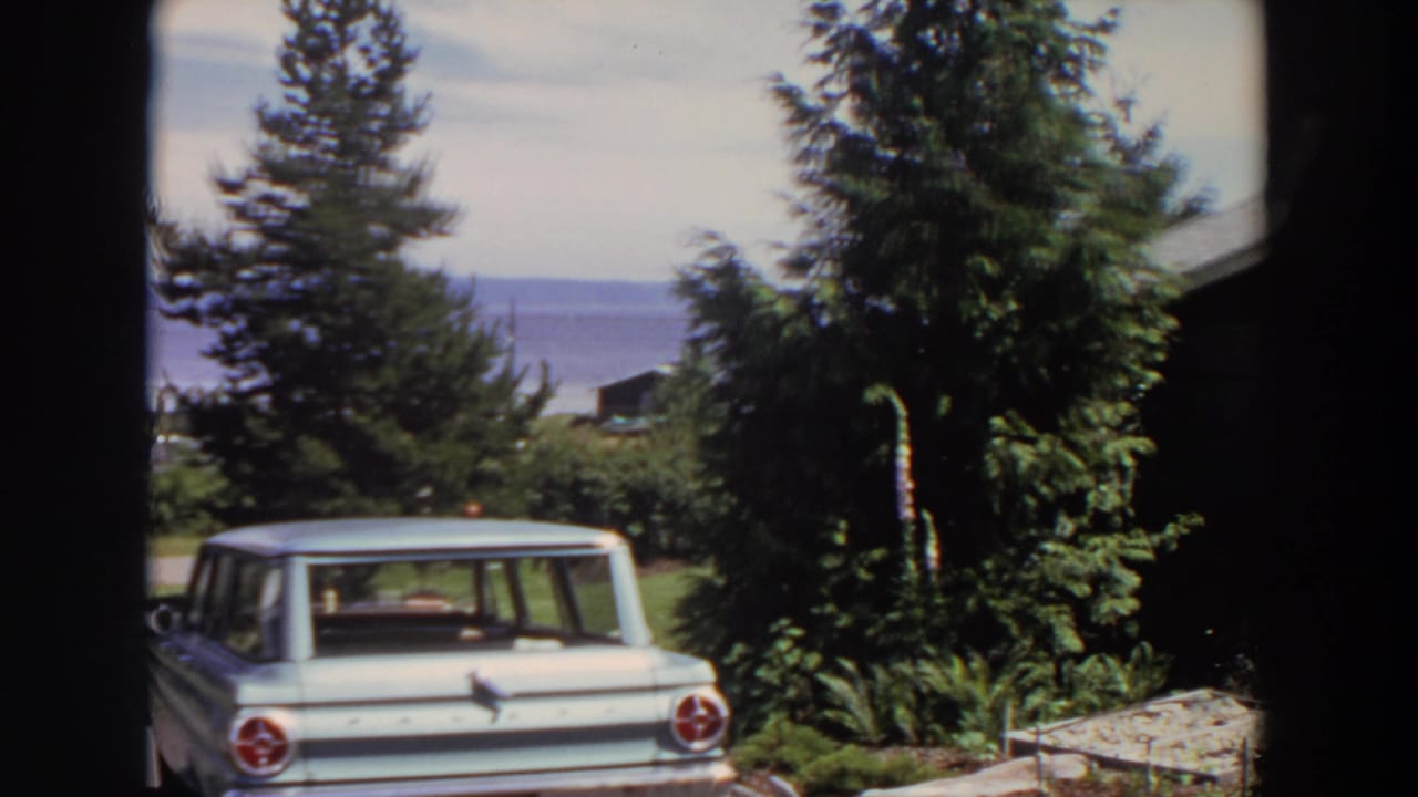 Late frame of the 1964 Washington pan showing a pale wagon, leafy yard plantings, conifers, road, distant water, and hills.