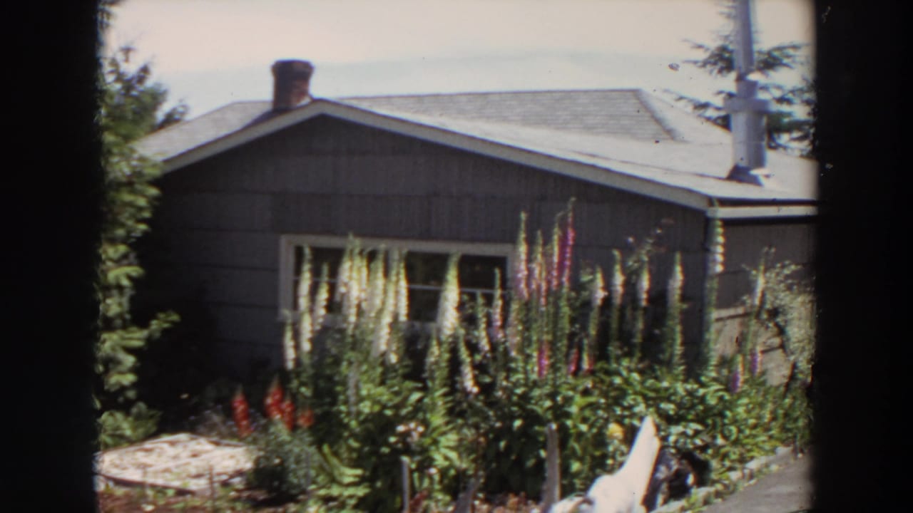 1964 Washington frame showing a grey residential exterior, flower garden, trees, paved edge, and partial dark window-like borders.