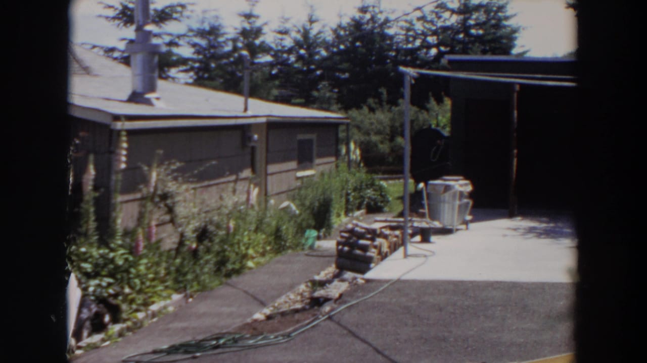 1964 Washington backyard frame with a paved area, coiled garden hose, stacked firewood, planted border, and the side of a modest house.