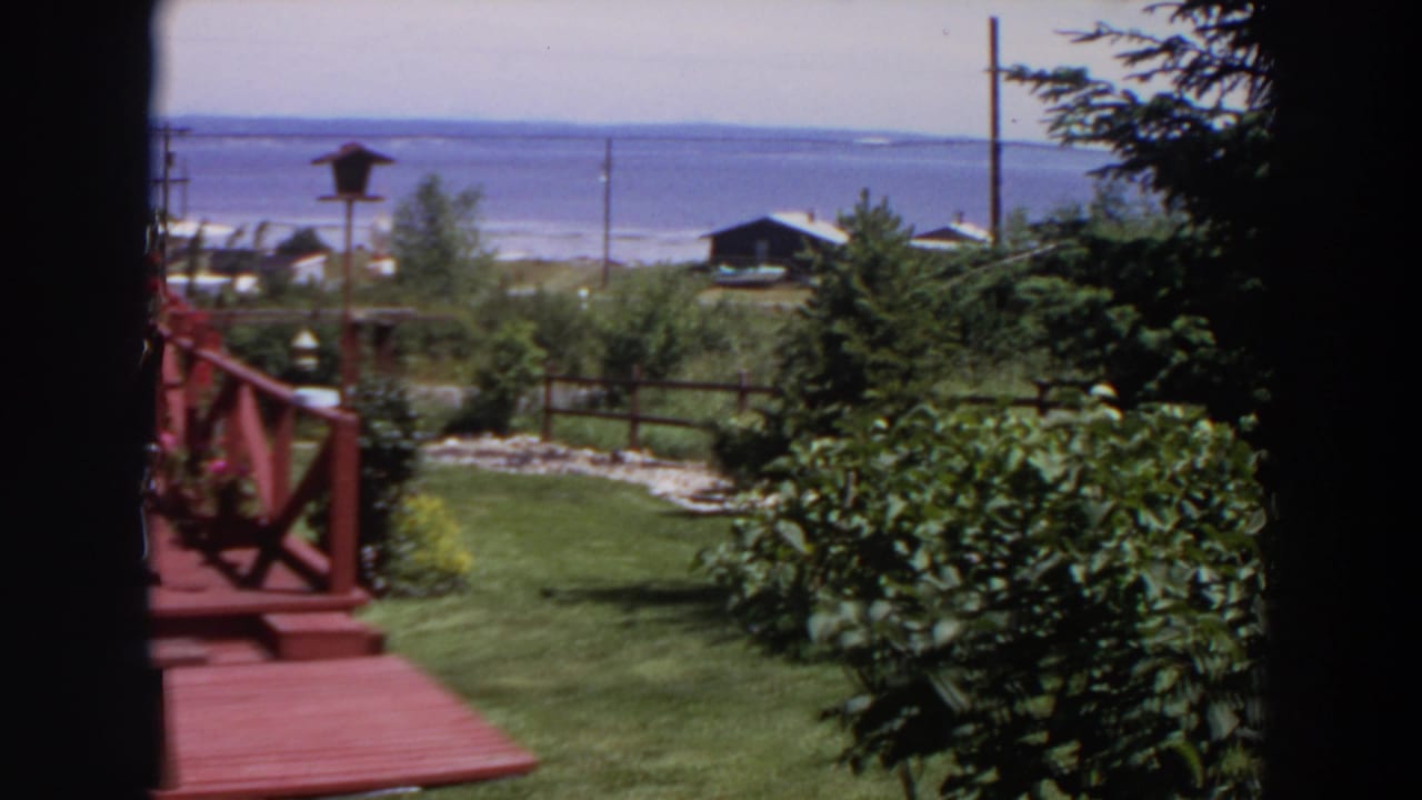 1964 Washington 8mm frame with part of a red wooden deck, garden shrubs, lawn, fence, trees, shoreline buildings, and water.