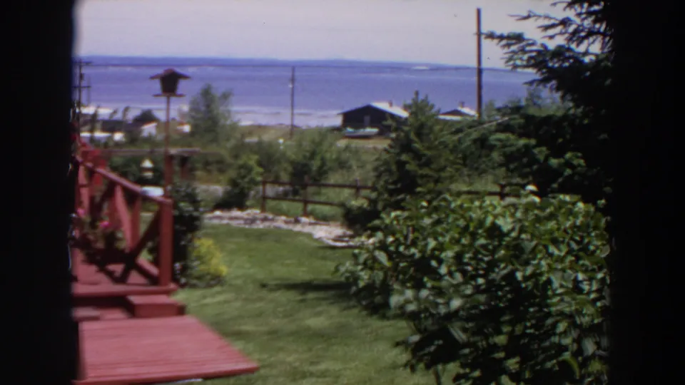 1964 Washington 8mm frame with part of a red wooden deck, garden shrubs, lawn, fence, trees, shoreline buildings, and water.