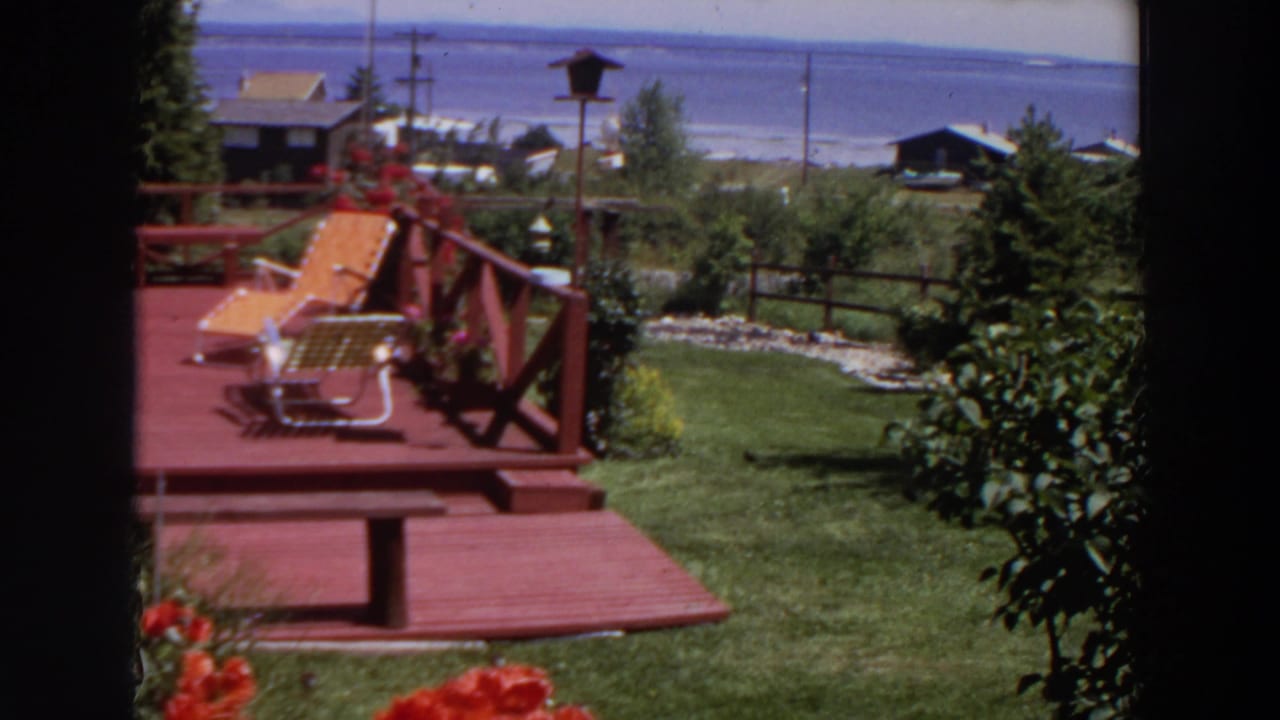 1964 Washington home-movie view of a red wooden deck with a lounge chair, garden plants, lawn, fence, small buildings, and water beyond.