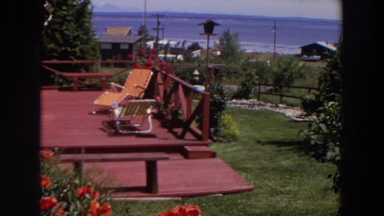1964 Washington home-movie view of a red deck with lounge chairs, garden flowers, lawn, nearby houses, and distant water.