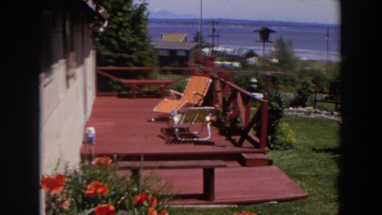1964 Washington yard scene with a red deck, two lounge chairs, birdhouse, garden shrubs, house wall, and water beyond.
